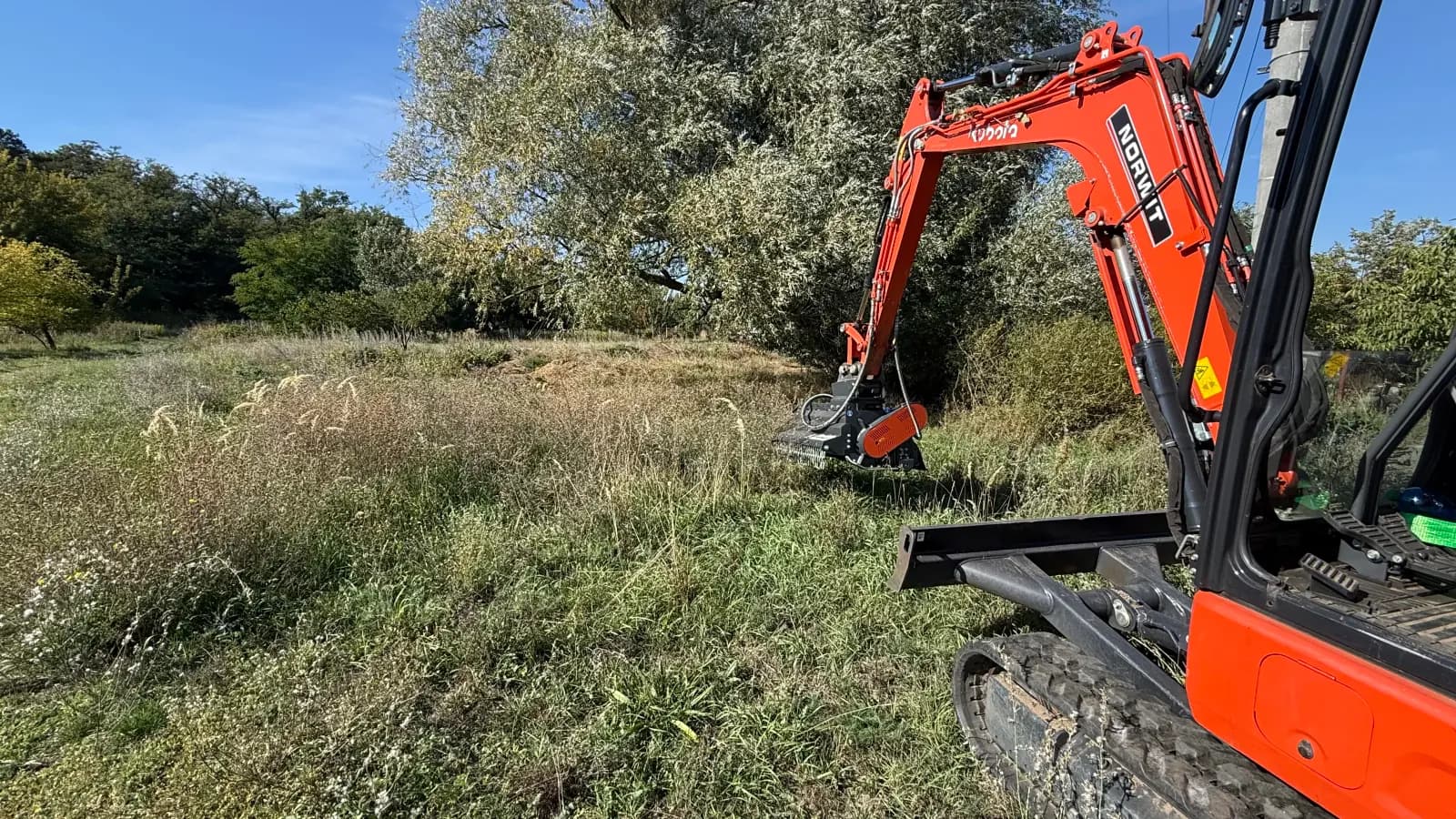 Minibager s mulčovacou hlavou medzi vysokou trávou a stromami v Malackách